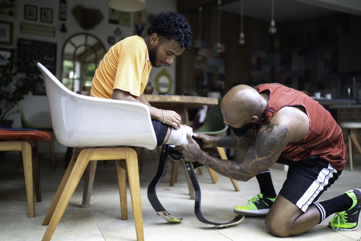 A Black father kneels to the floor while helping his teenage son fit on a prosthetic running blade